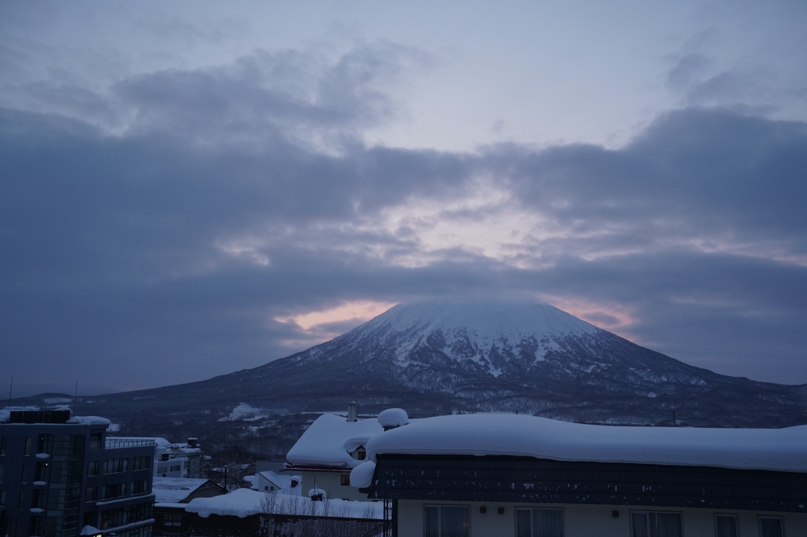 A snow-capped volcanic mountain under heavy cloud at dusk, seen over snow-covered rooftops