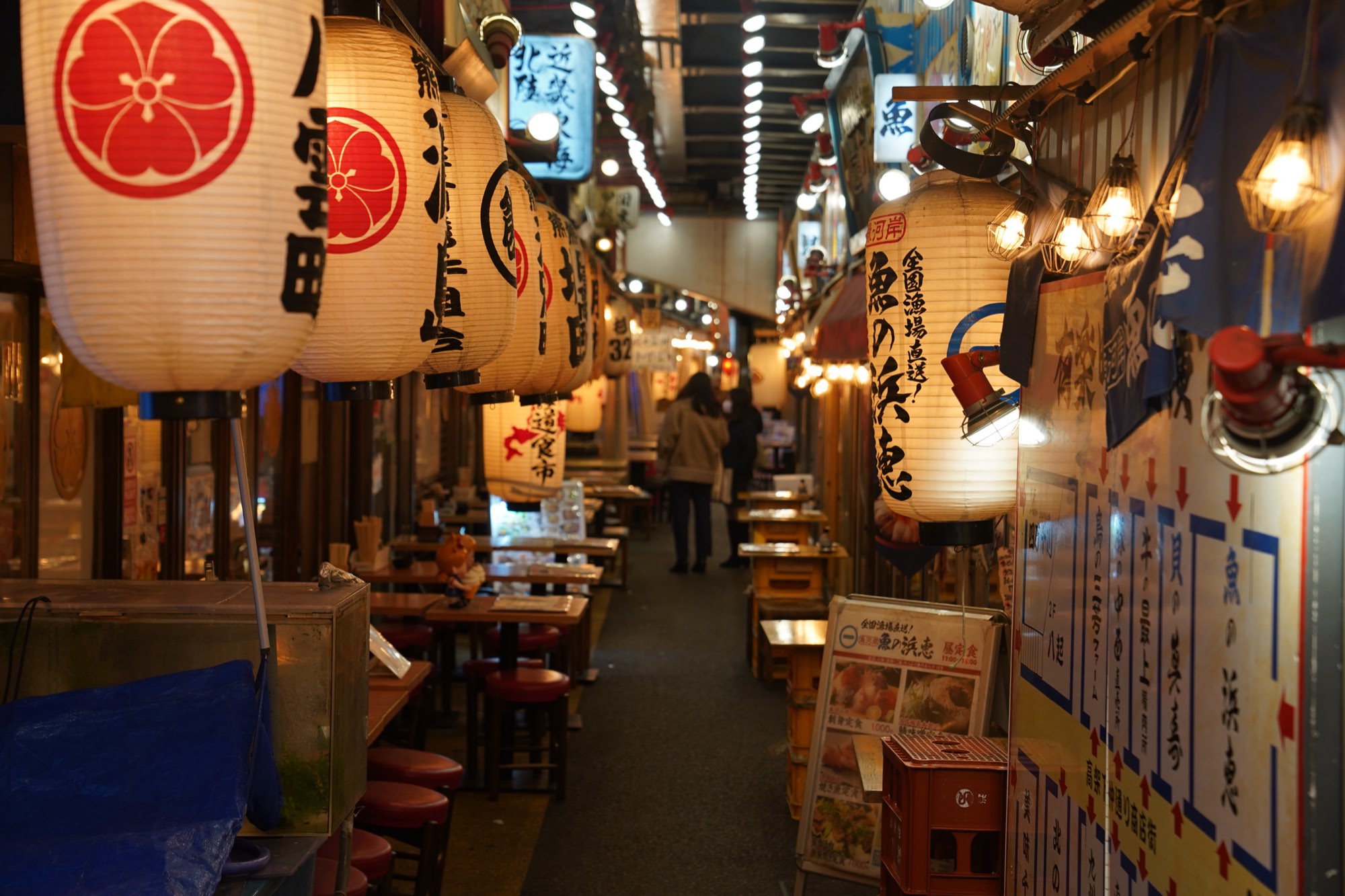 A narrow restaurant alley in Tokyo at night, lined with large illuminated paper lanterns and signs, its walkway receding into the distance